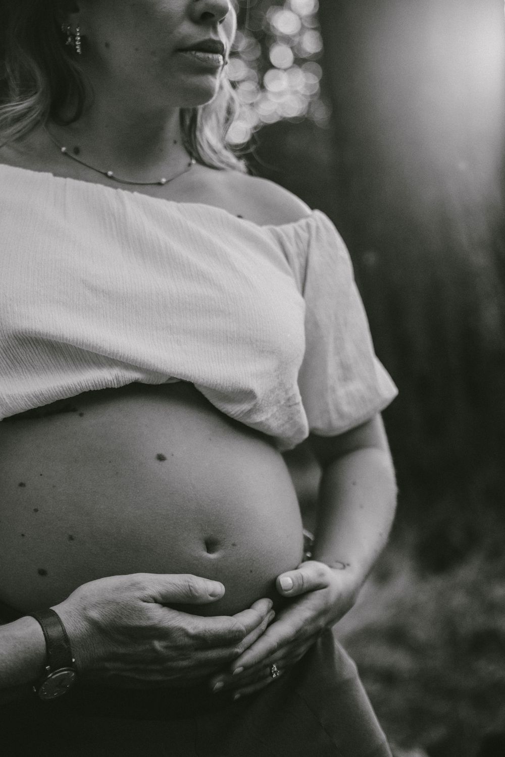 Black and white close-up of pregnant woman gently cradling her bare baby bump in soft natural light outdoors.