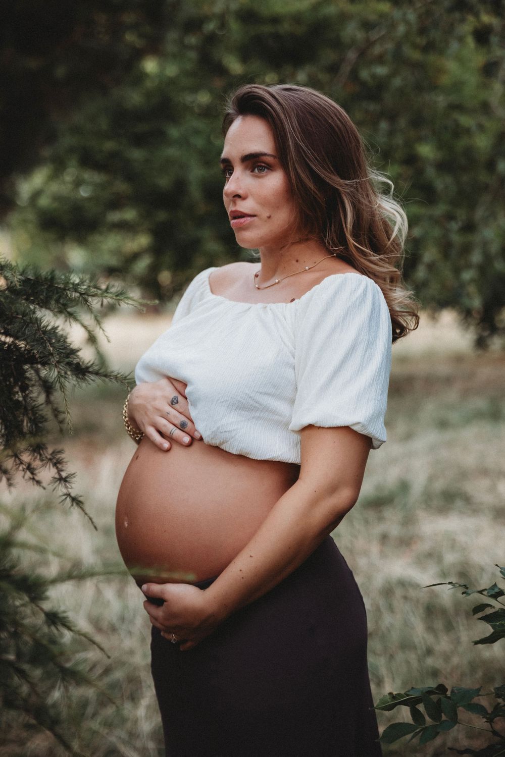 Pregnant woman in white off-shoulder top holds baby bump while standing among green trees in natural outdoor setting.