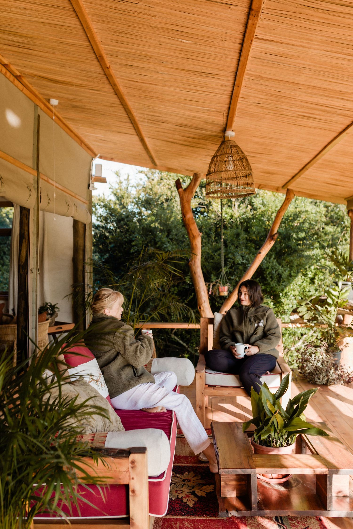 Two women relaxing on a cozy outdoor porch with plants, wicker pendant light, and bamboo ceiling.