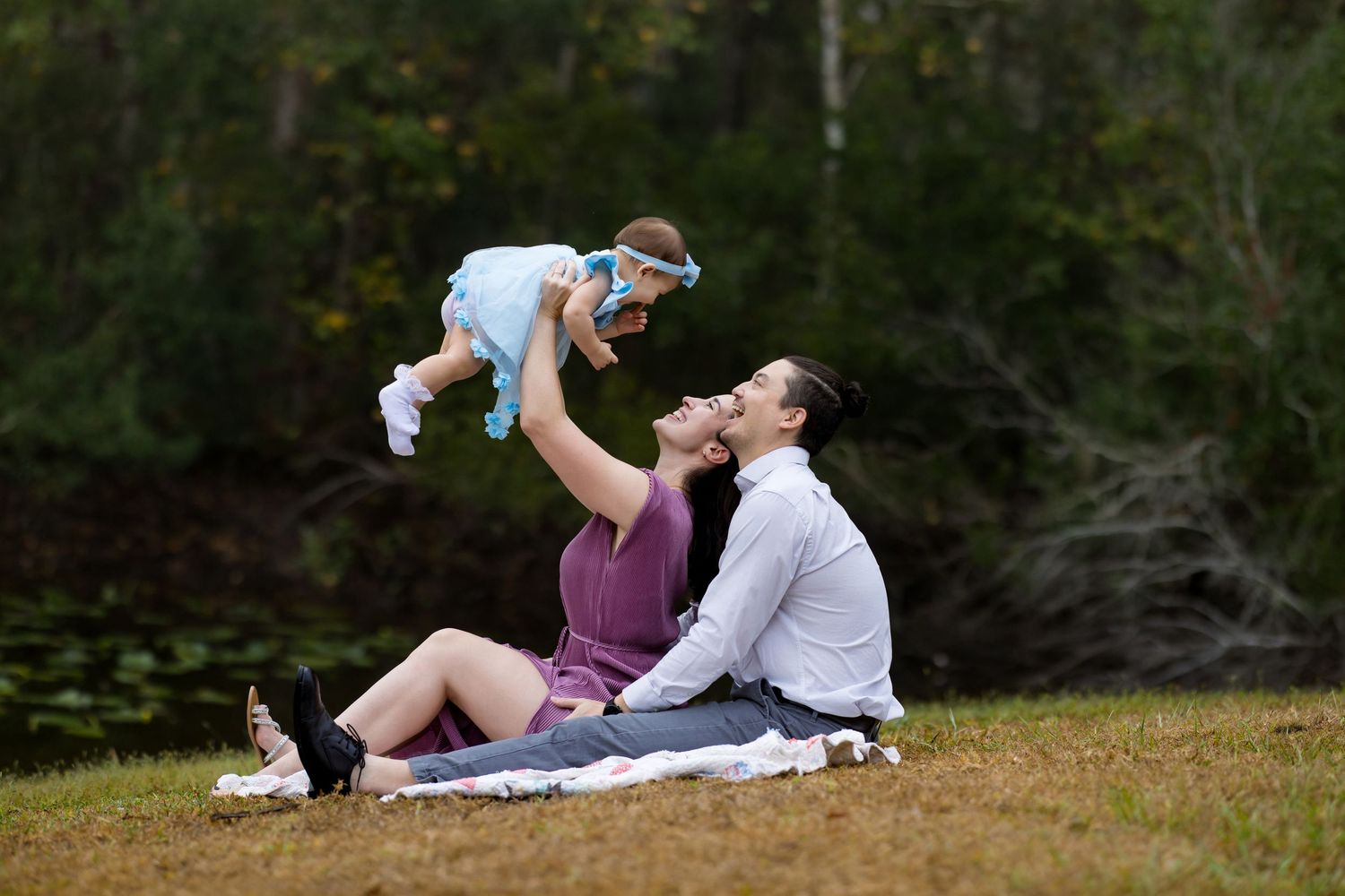 A couple sits on a blanket outdoors, joyfully lifting their baby girl in a blue dress against a lush green backdrop.