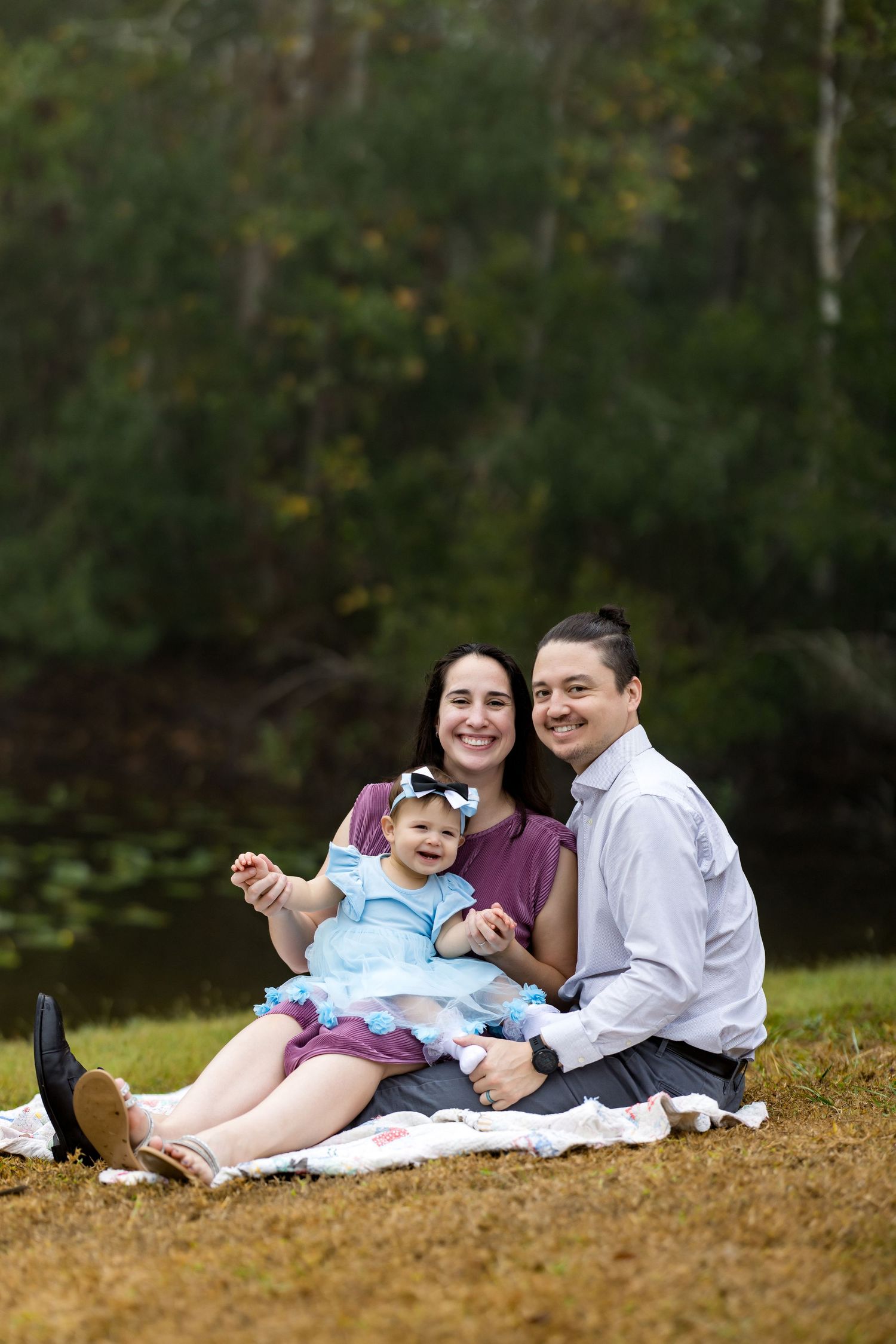 Family of three sitting on blanket outdoors, parents smiling with baby girl in blue dress, green trees background.