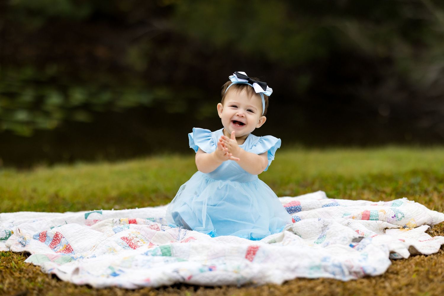 A happy baby girl in a blue dress sits on a quilt blanket outdoors, clapping her hands and smiling.