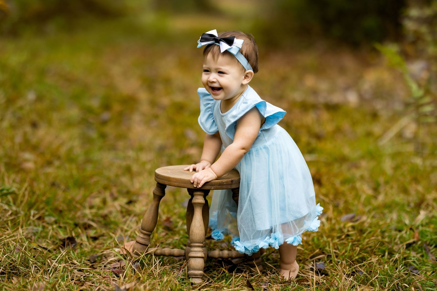 A laughing baby girl in a blue dress and bow headband stands outdoors holding a small wooden stool.