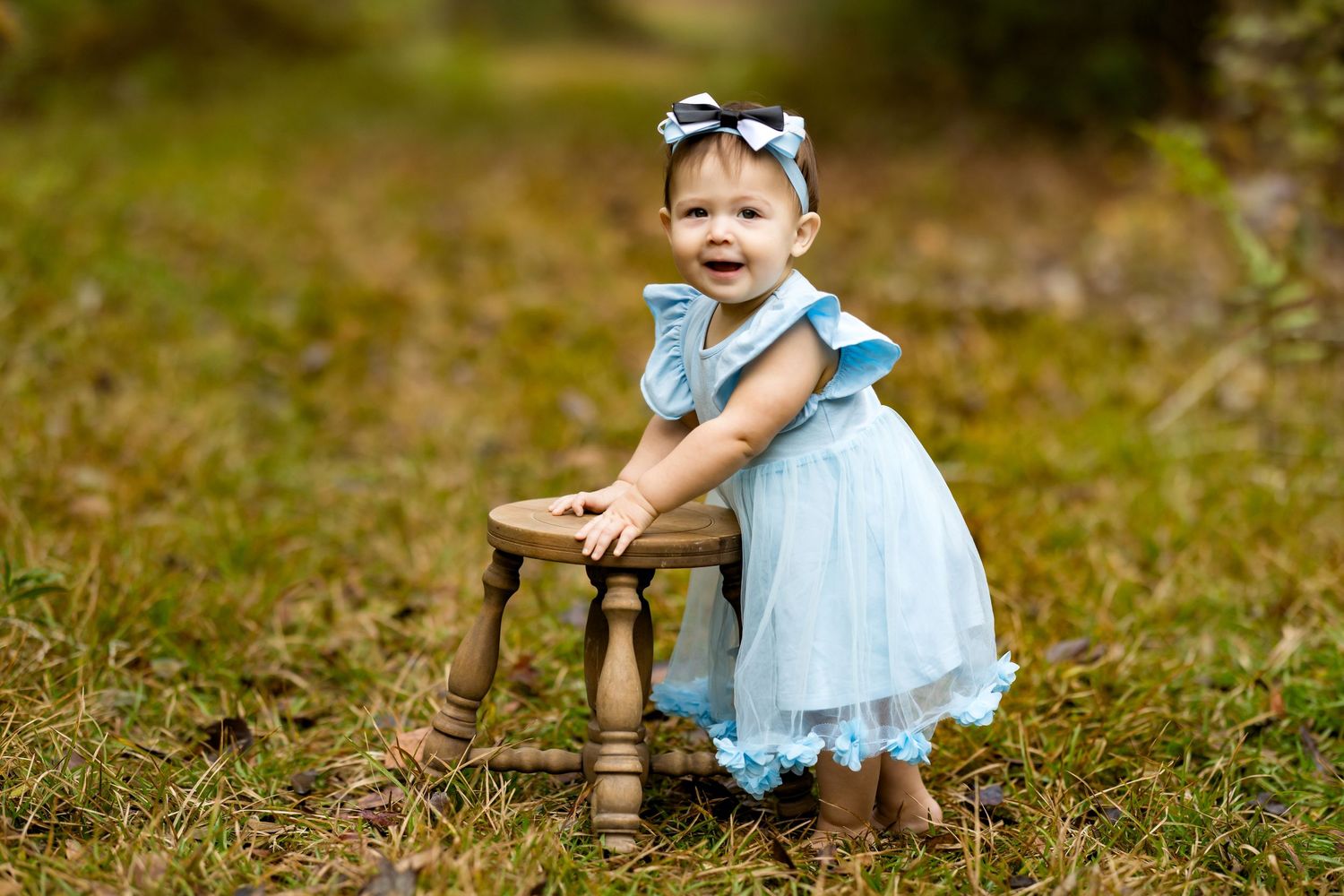 Toddler girl in blue dress with bow headband standing by wooden stool outdoors in garden setting.