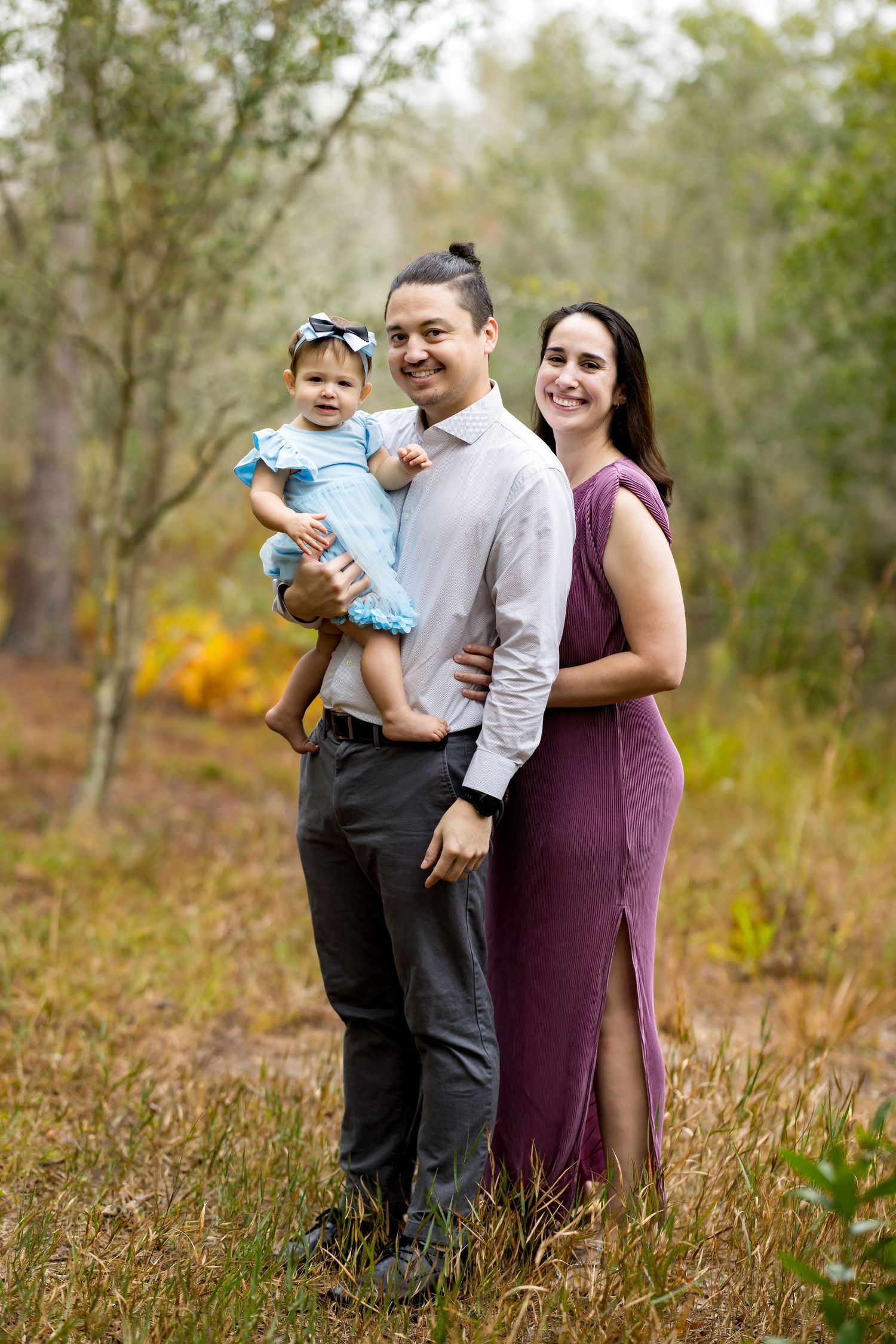 A young family poses outdoors in a forest, man holding baby in blue dress, woman in purple gown, smiling together.
