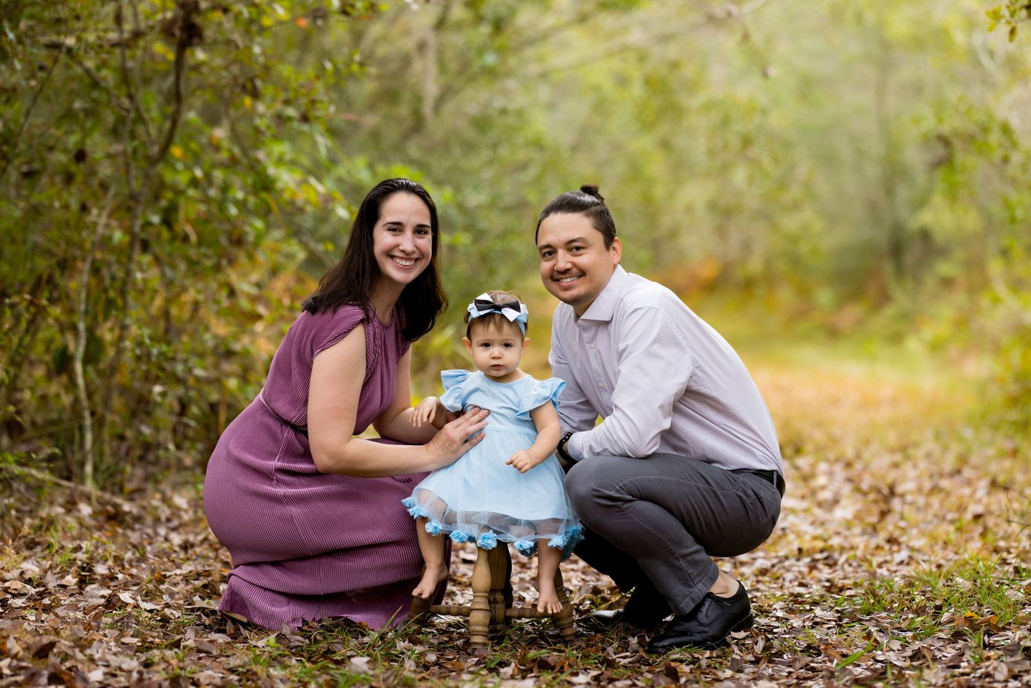Family of three posing outdoors on a leaf-covered path, parents kneeling beside their baby girl in a blue dress.