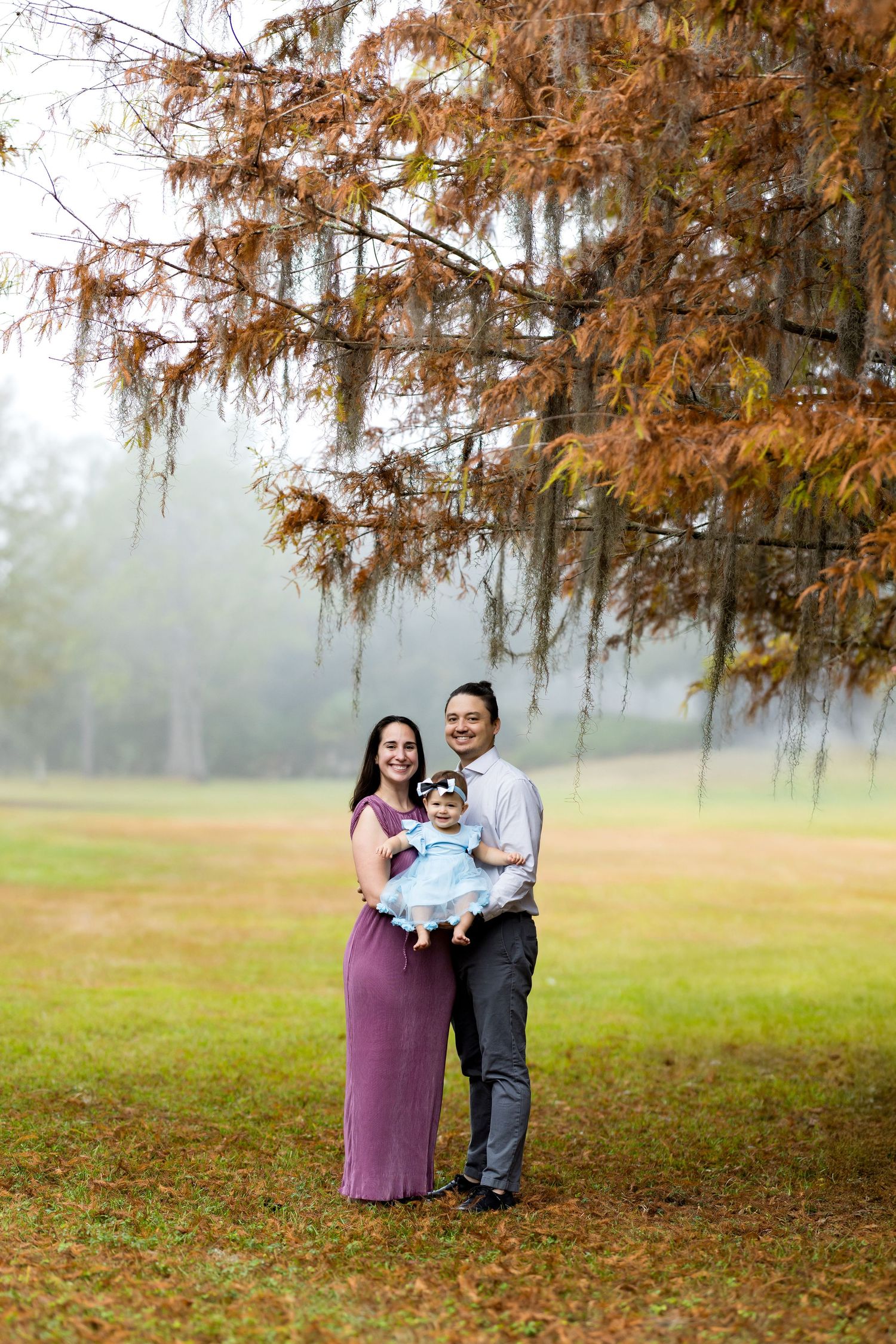 Family of three posing outdoors under autumn tree in misty park, mother in purple dress holding baby girl in blue dress.
