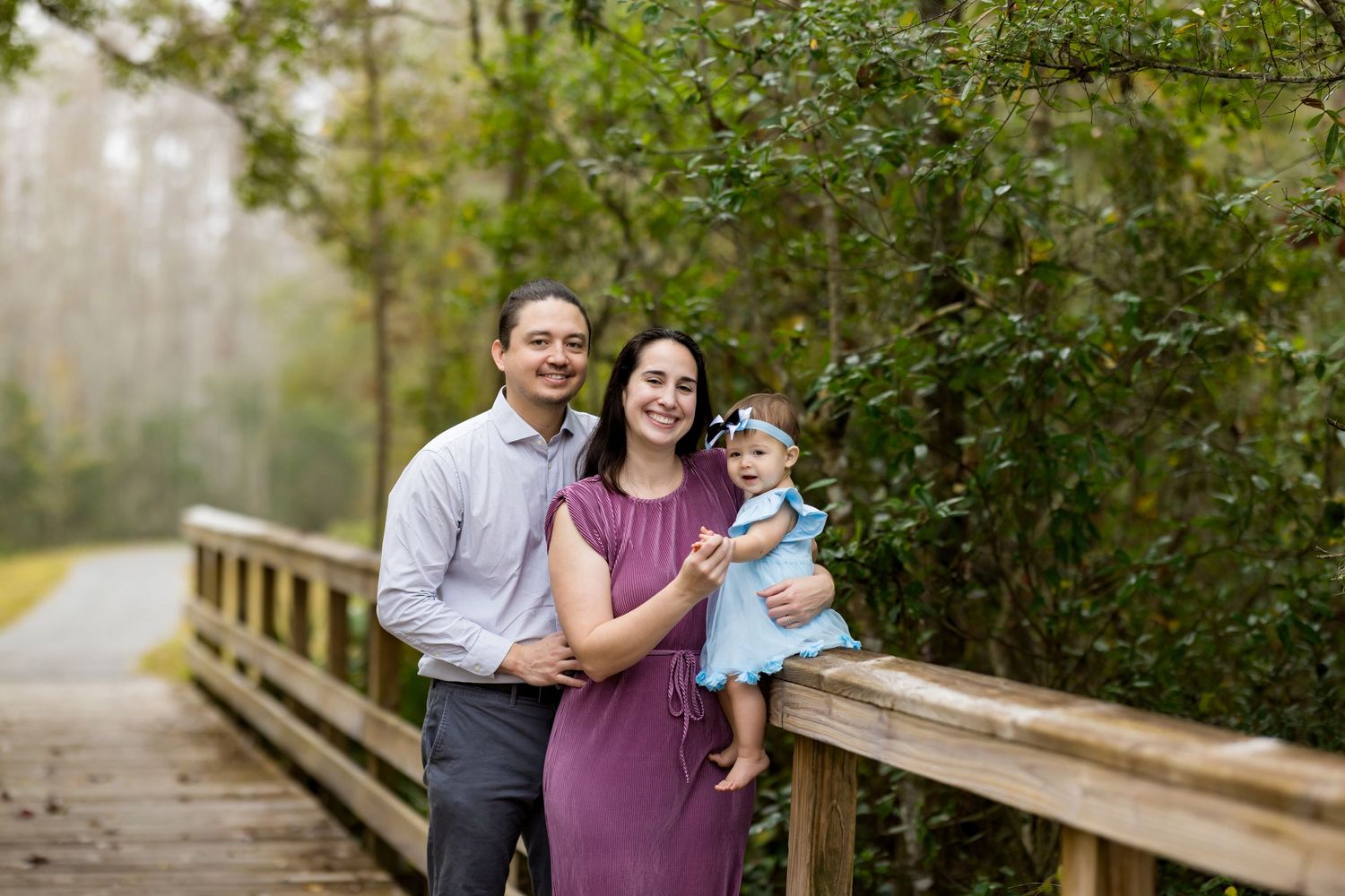 A smiling family of three poses on a wooden bridge surrounded by lush greenery, with parents holding their baby girl.
