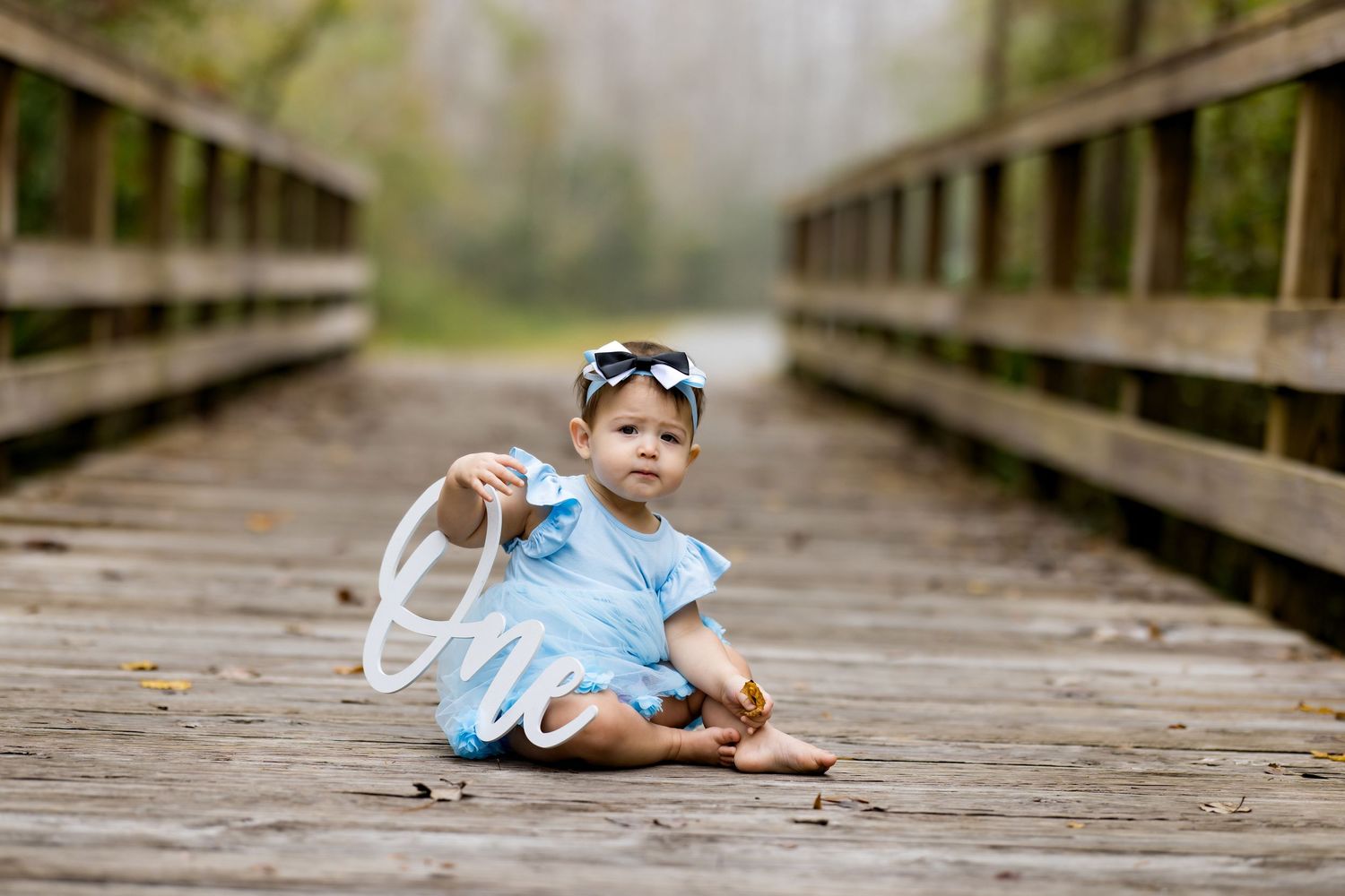 Baby girl in blue dress sits on wooden bridge holding white letter, wearing bow headband, surrounded by nature.