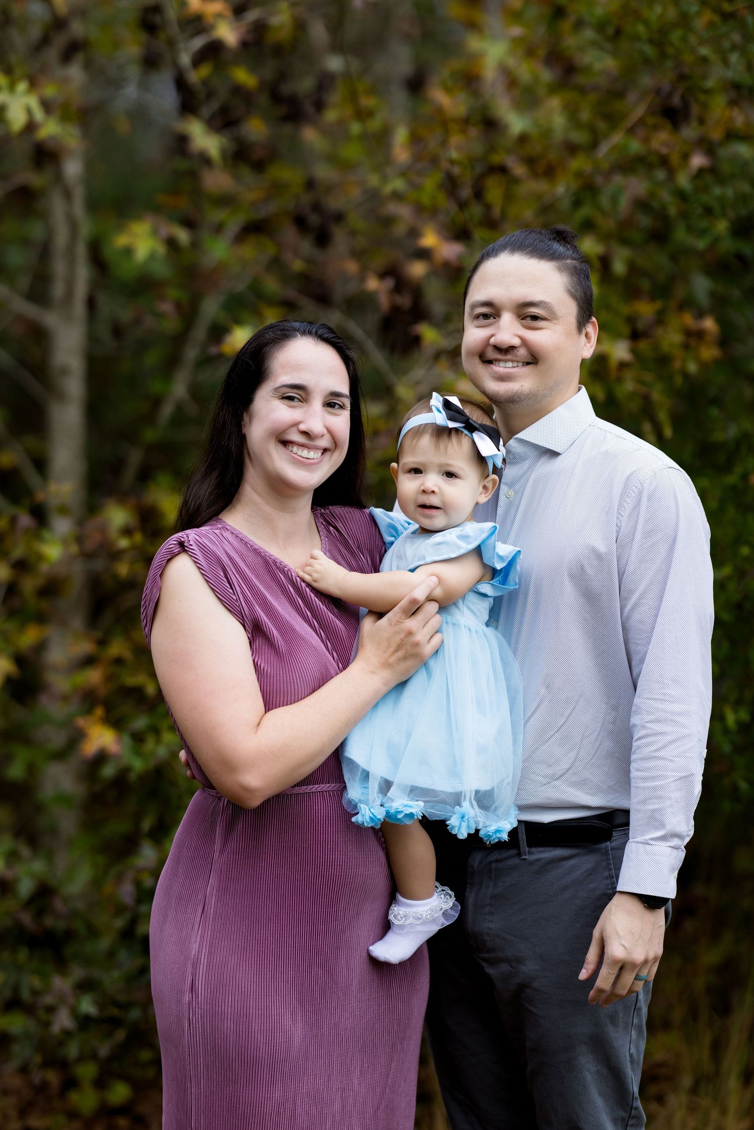 Smiling family portrait with mother in purple dress, father, and baby girl in blue dress outdoors.