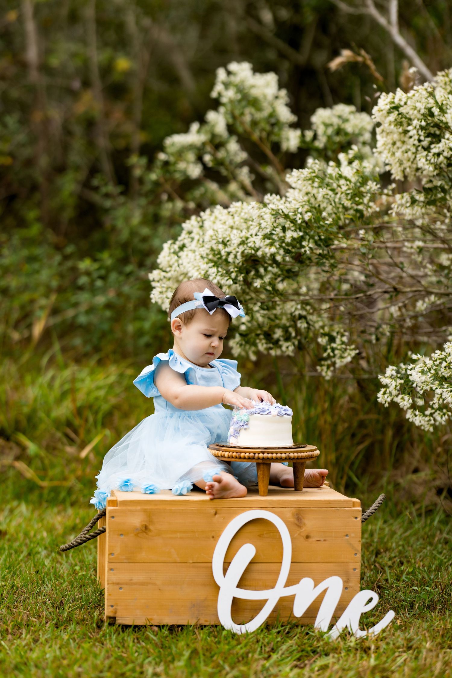 Baby girl in blue dress smashes her first birthday cake outdoors, with a wooden crate displaying the word One.