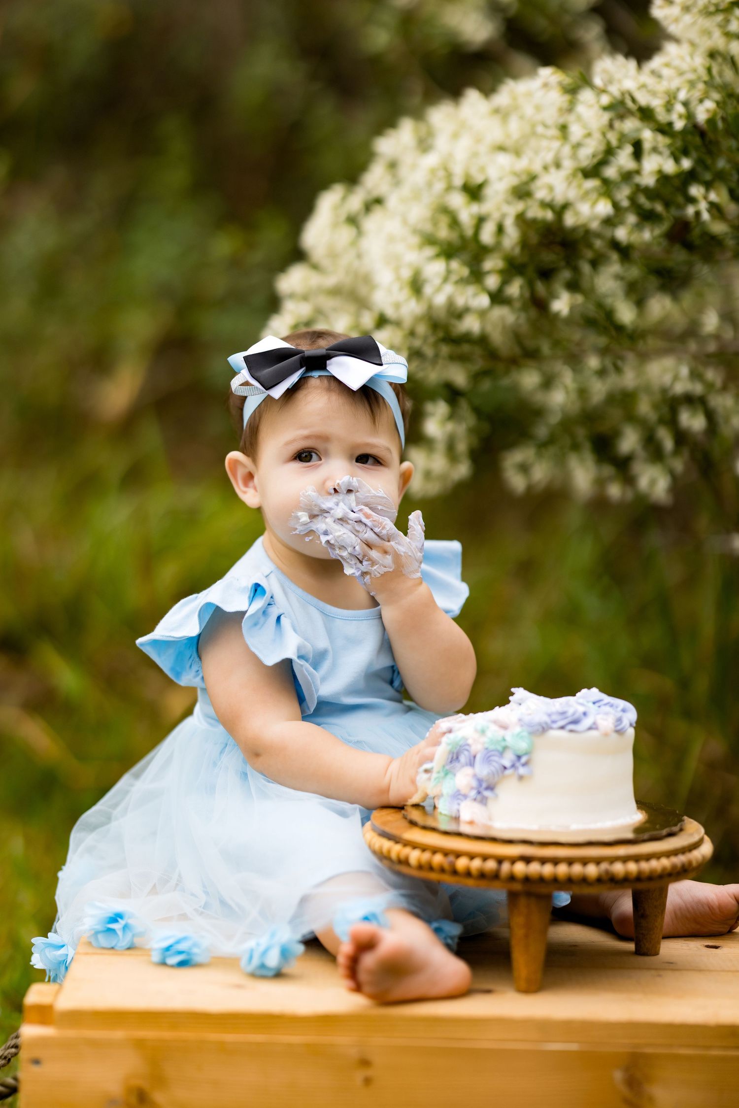Baby girl in blue dress eating cake with hands at outdoor first birthday cake smash photo session.