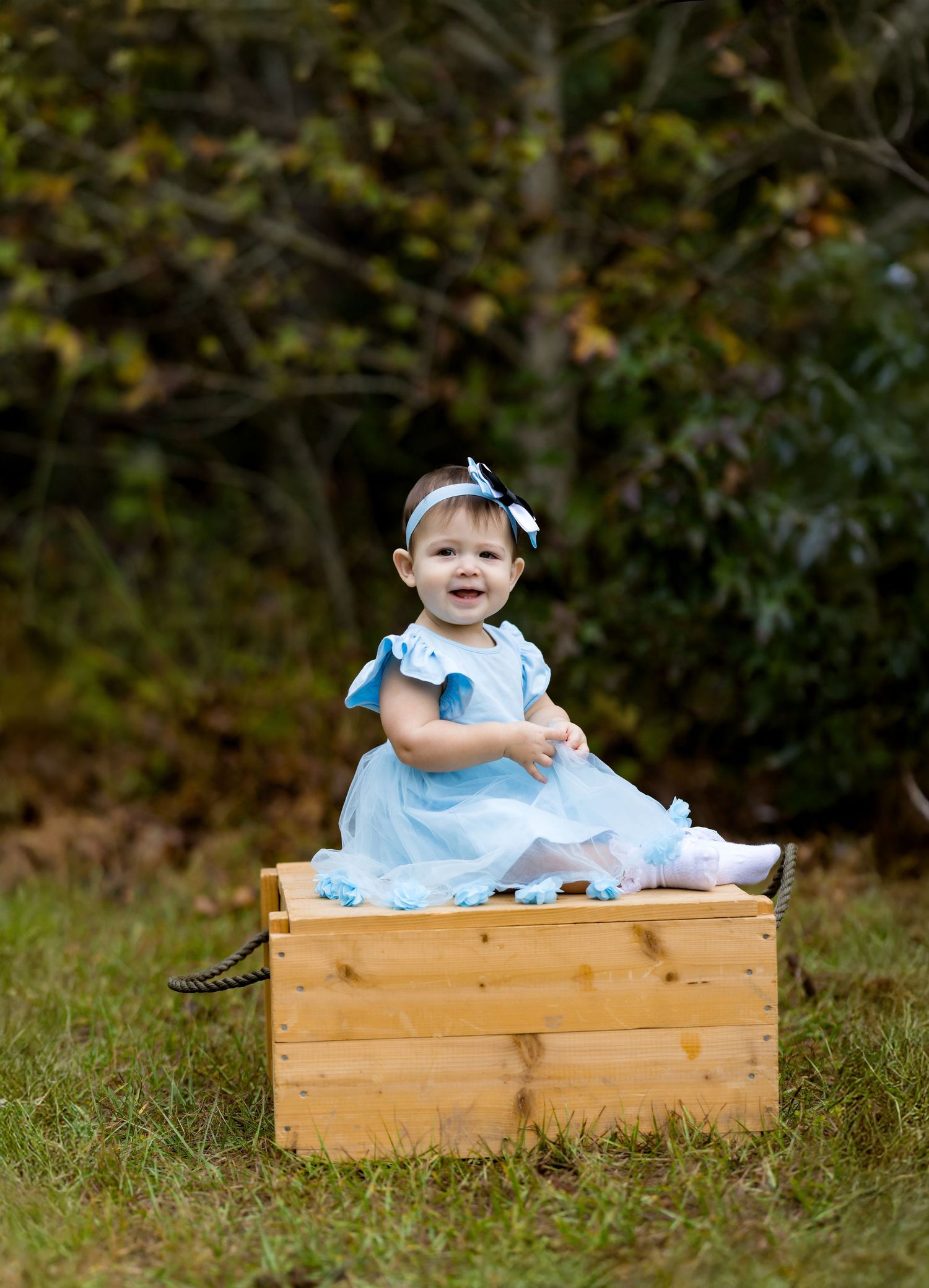 A smiling baby girl in a light blue dress and headband sits on a wooden crate outdoors among green trees.