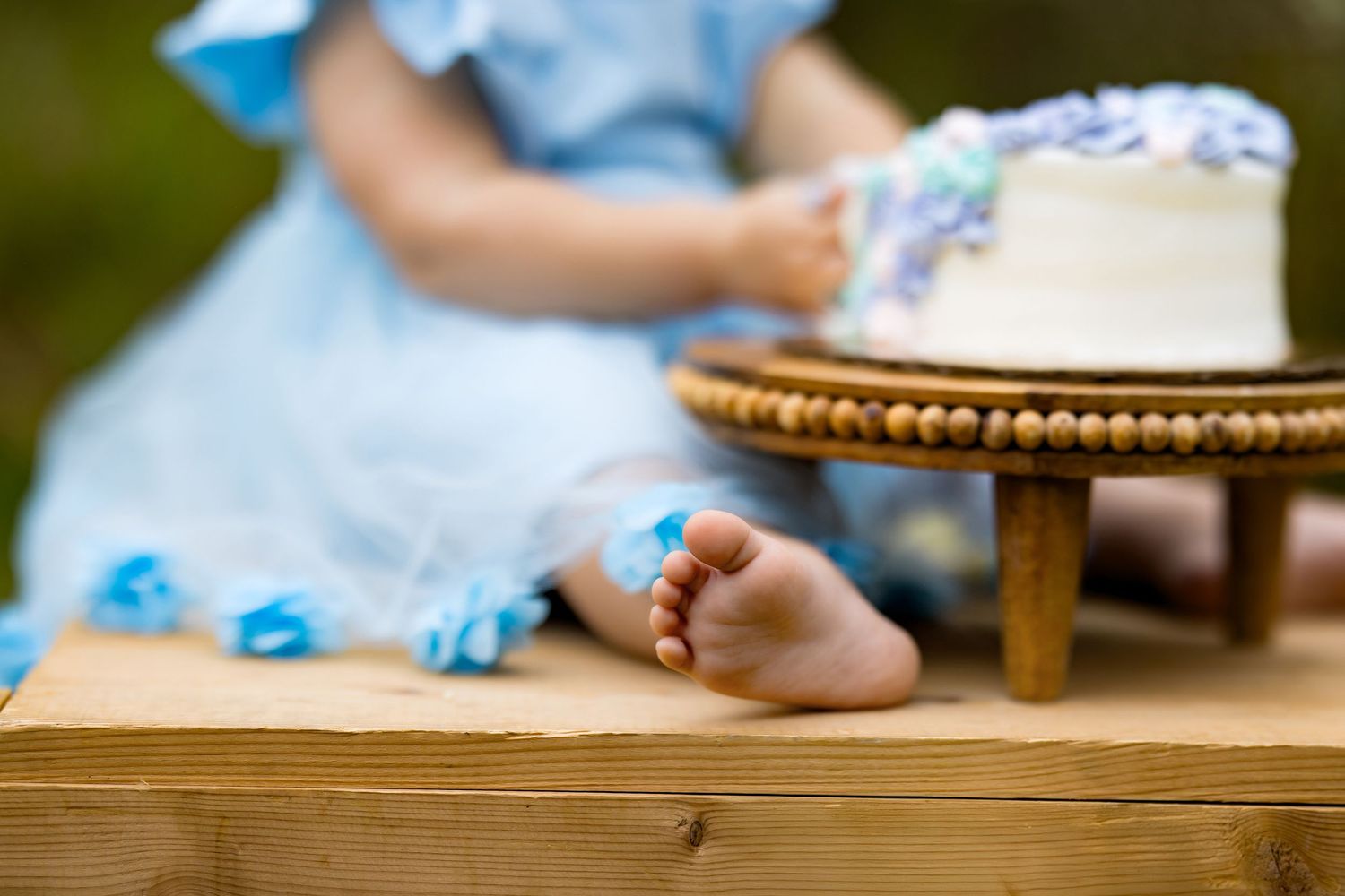 Baby in blue dress sitting on wooden bench beside white cake on stand with blue floral decorations.