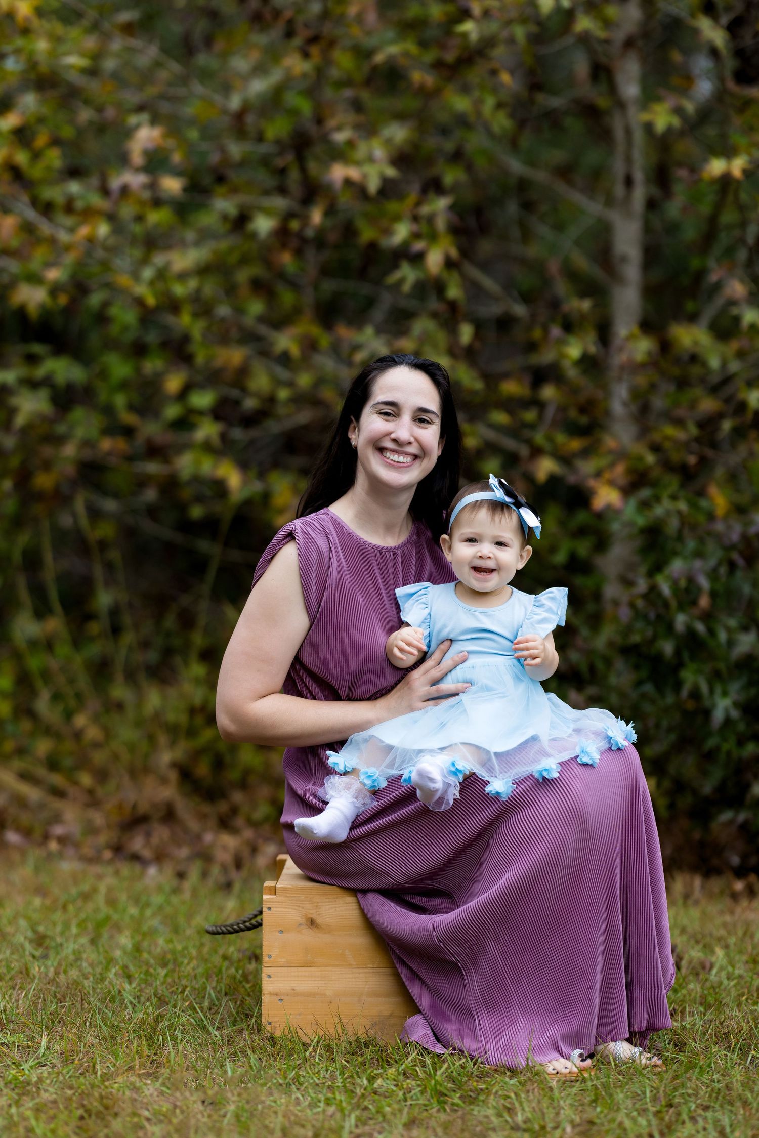 A smiling woman in a purple dress sits outdoors holding a happy baby girl in a blue dress with floral trim.