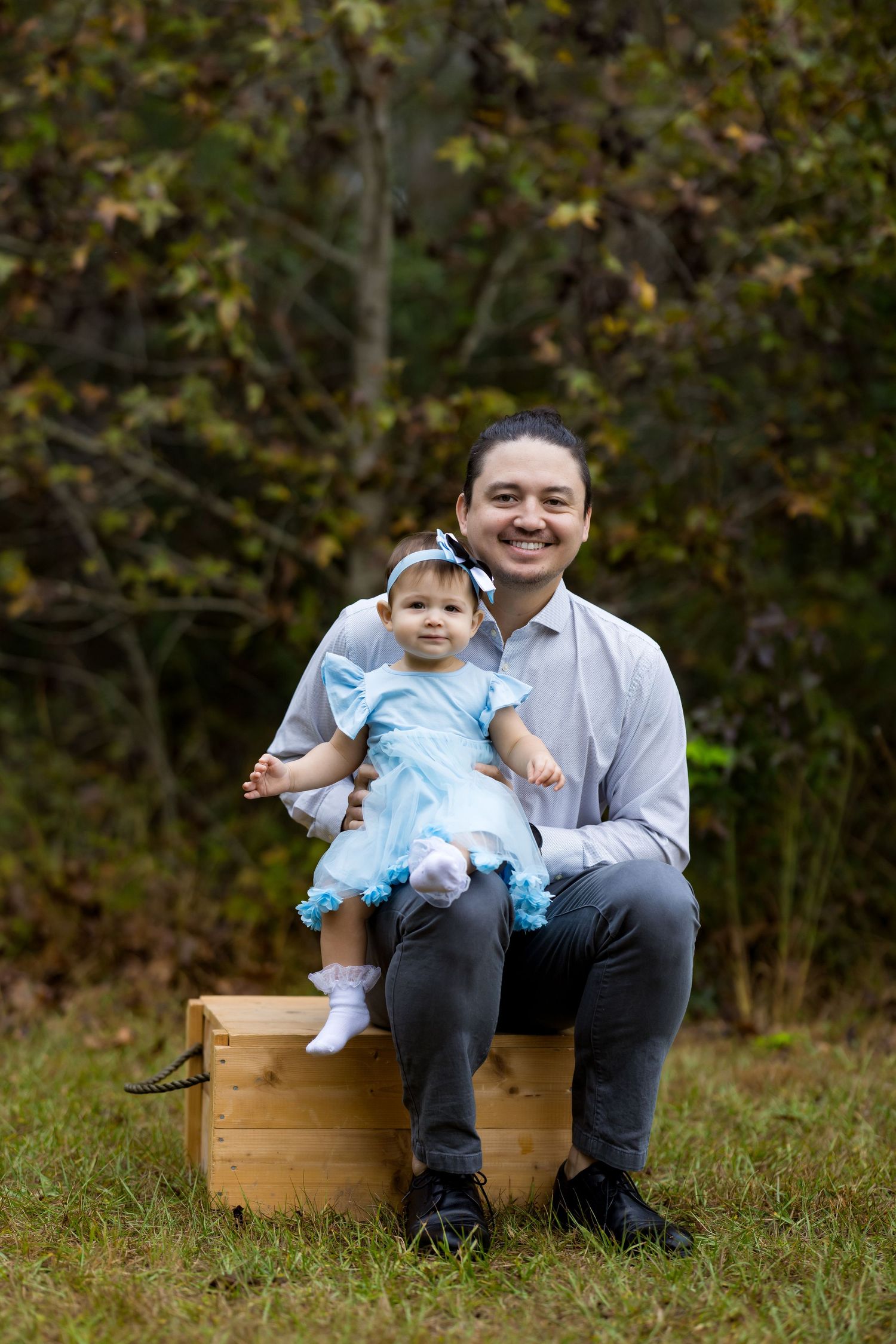 A smiling man sits on a wooden crate outdoors holding a baby girl in a blue dress, surrounded by autumn foliage.