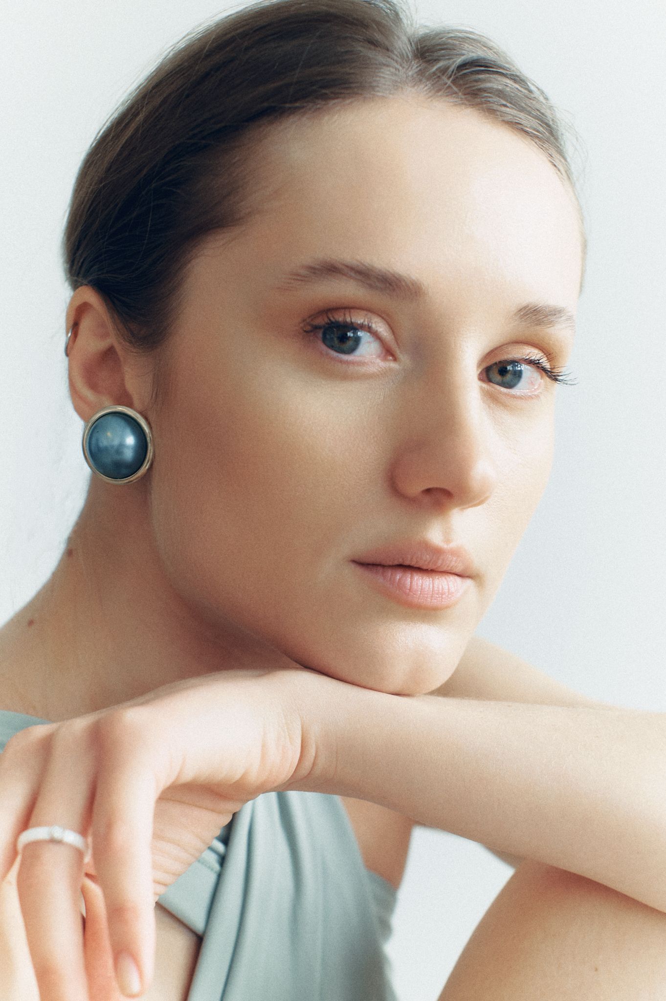 Young woman with blue eyes and dark pearl earring poses thoughtfully in sage green top against white background.