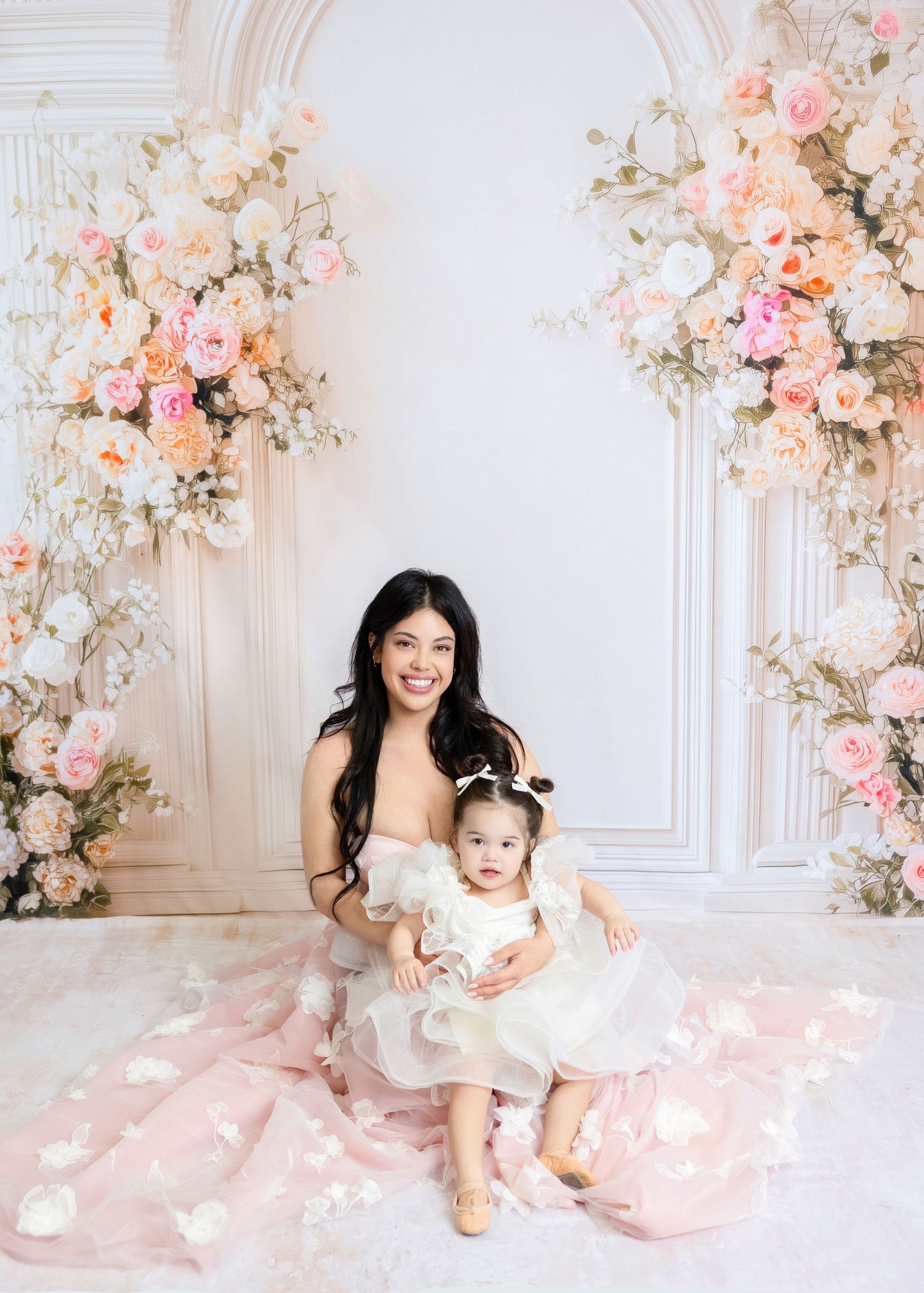 A woman and baby girl in white dresses pose together before a floral arch with pink and peach roses.