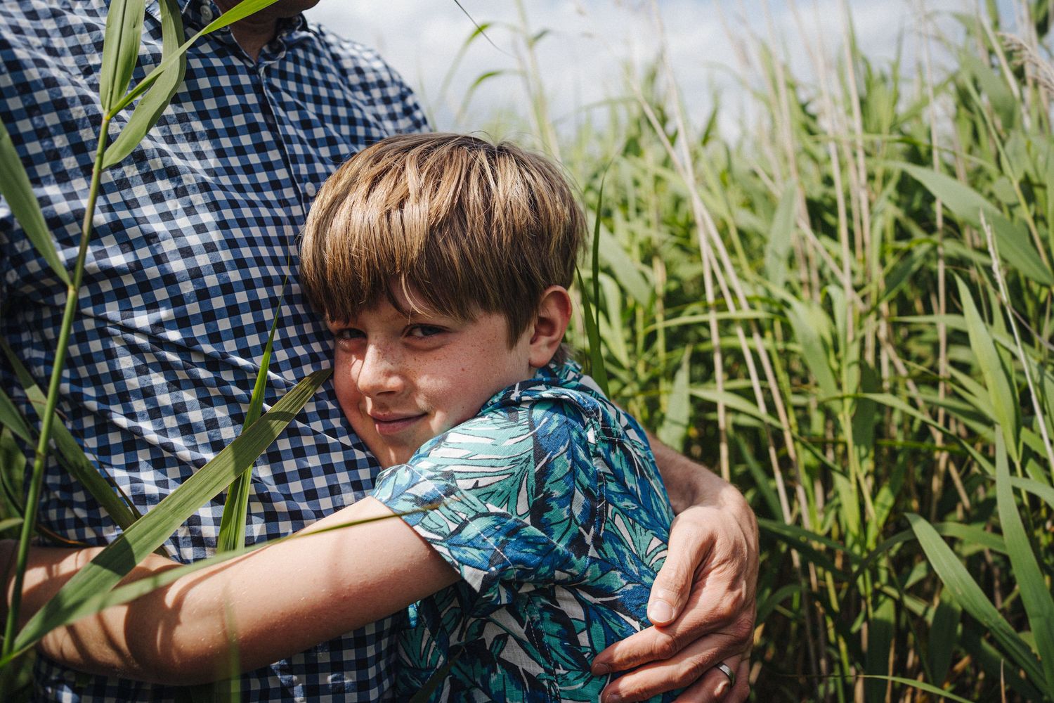 A young boy hugging an adult outdoors, smiling among tall green grass on a sunny day.