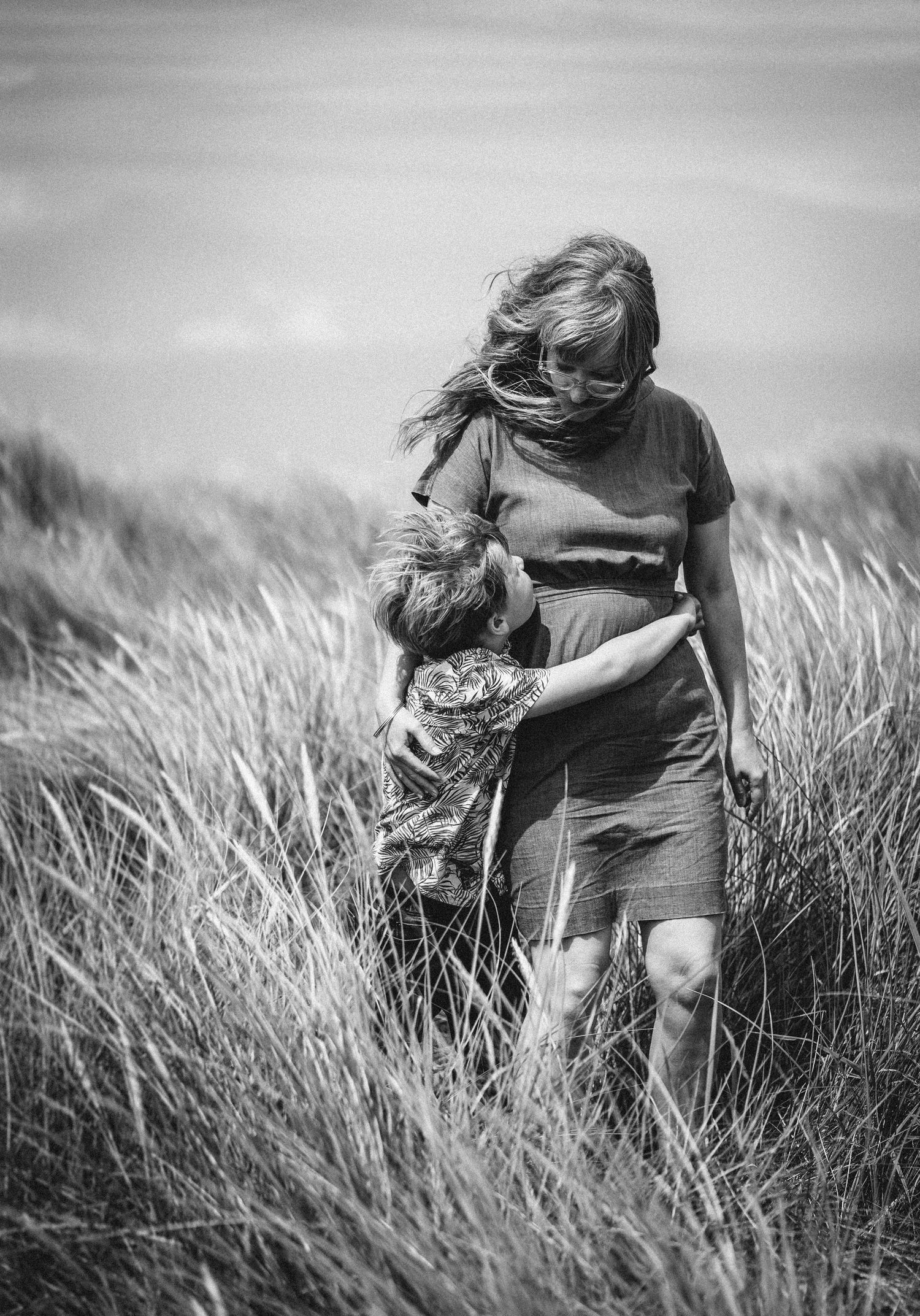 Black and white photo of a mother and child embracing in a grassy field, with wind blowing through her hair.