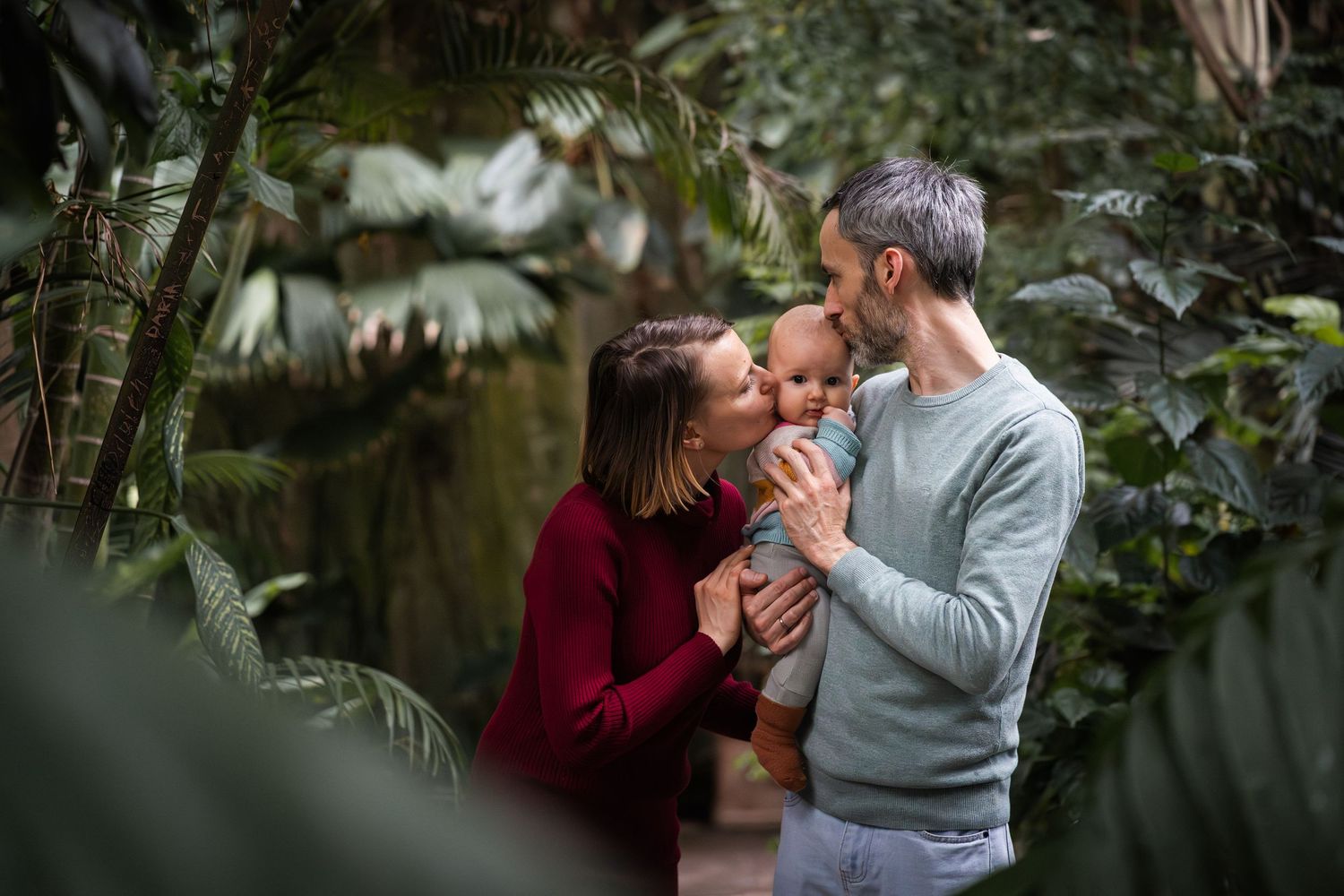 A couple shares a tender moment kissing their baby amid lush tropical greenery in a botanical garden.