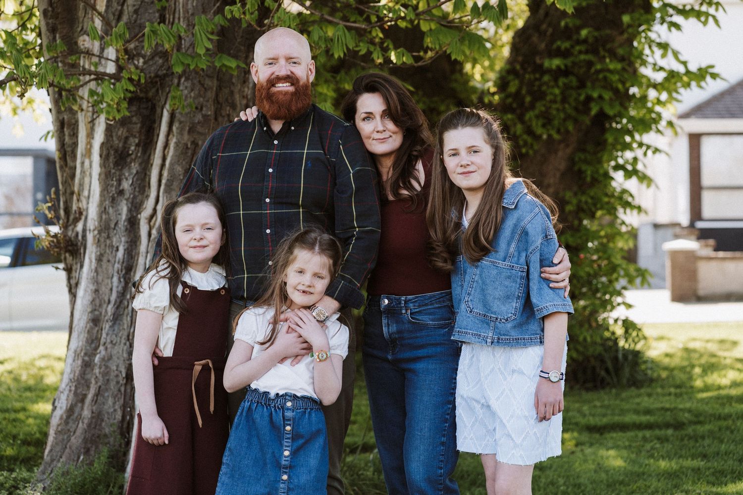 Family of five smiling outdoors under a tree, parents with three daughters posing together on a green lawn.