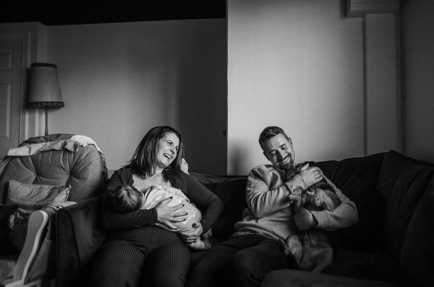 Black and white photo of a family with a newborn baby sitting on a couch, parents smiling warmly in a cozy living room.