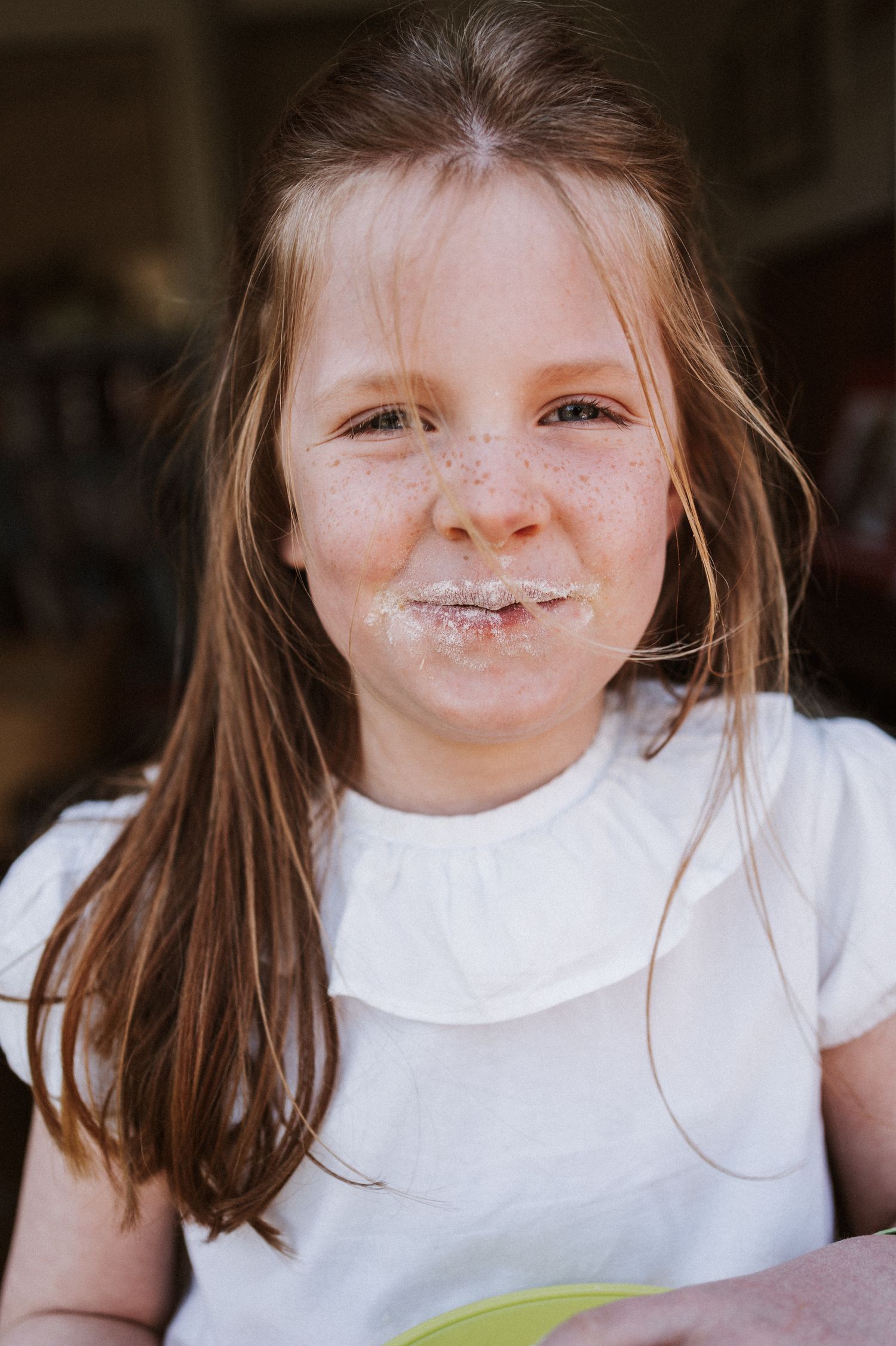 Young freckled girl with messy face smiling after eating cake, wearing white top with long brown hair.