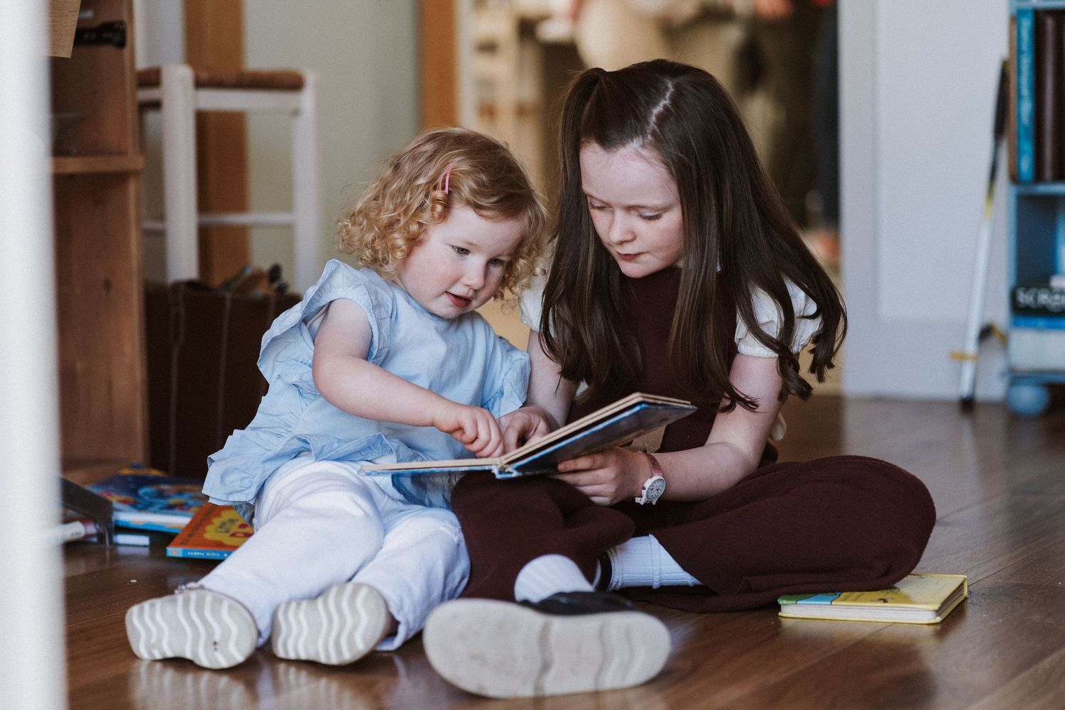 Two young girls sitting on hardwood floor reading a book together in a cozy home setting.