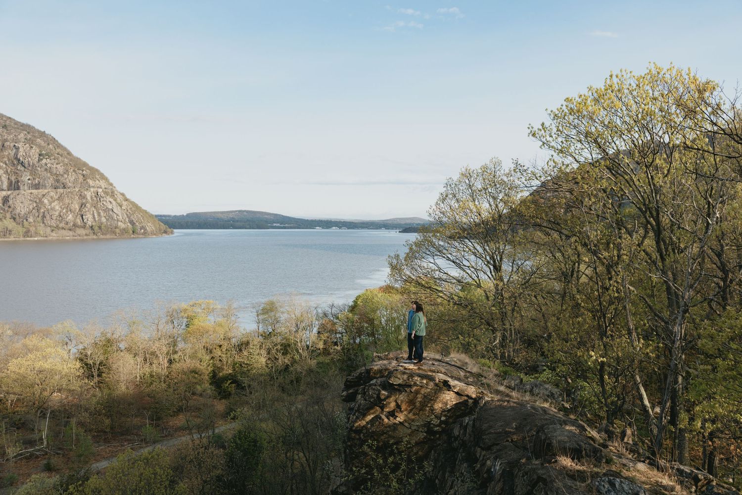 A hiker stands on a rocky overlook surrounded by trees with a scenic river valley and mountain backdrop.