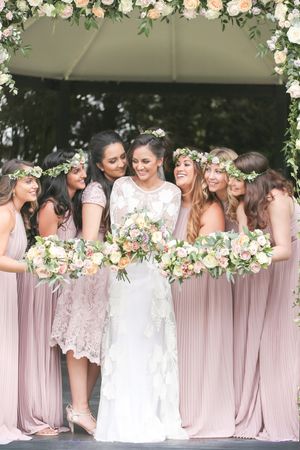 Bride in white lace gown with bridesmaids in blush pink dresses and floral crowns holding bouquets at outdoor wedding.