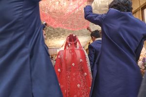 Traditional Chinese wedding ceremony with bride in red veil held by attendants in blue suits.