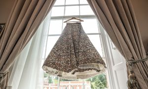 Floral patterned wedding dress hanging on a wooden hanger in front of a bright bay window with sheer curtains.