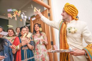 Groom in white sherwani and mustard turban tosses flower petals at Indian wedding ceremony with guests celebrating.