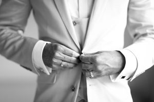 Black and white photo of a groom buttoning his suit jacket, wearing a ring on his finger.