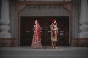 Sikh bride and groom in traditional red and gold wedding attire standing apart before ornate wooden doors.