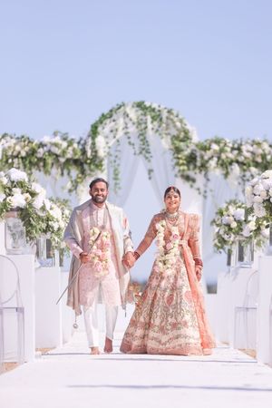 Indian bride in pink lehenga and groom in ivory sherwani walk down floral aisle at outdoor wedding ceremony.