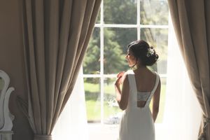 Elegant bride in backless white gown gazes out a sunlit window with flowing curtains on wedding day.