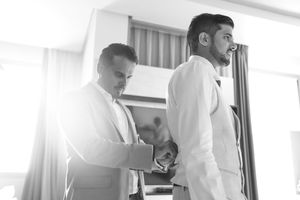 Two men in suits helping each other get dressed before a wedding ceremony in a bright room.