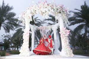 Bride in red lehenga posing under floral mandap with white flowers and palm trees at outdoor Indian wedding.