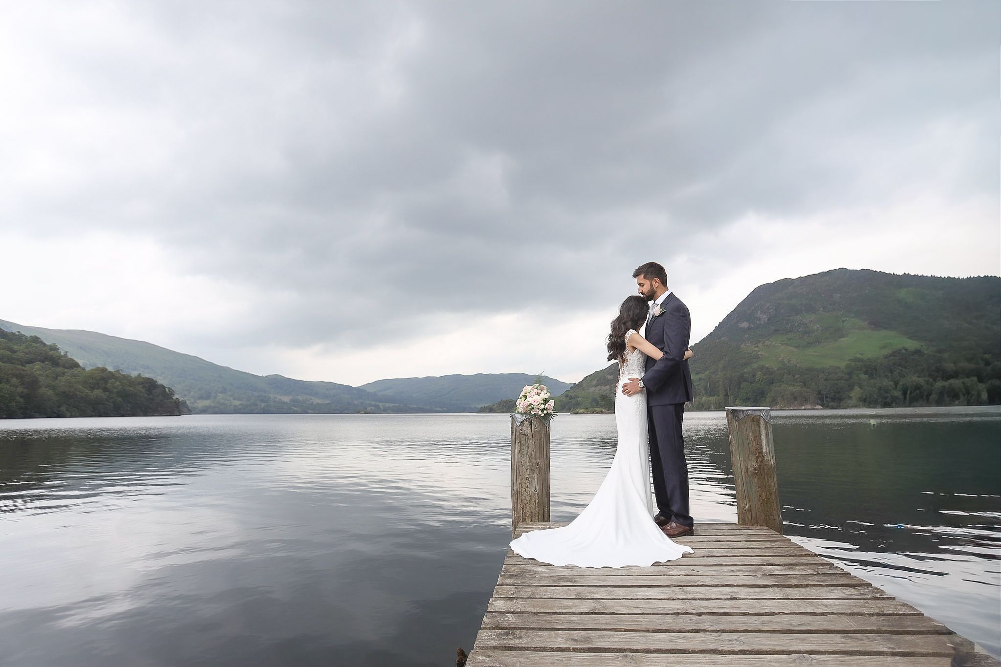 Bride and groom share a romantic moment on a wooden dock overlooking a serene lake with misty mountains.