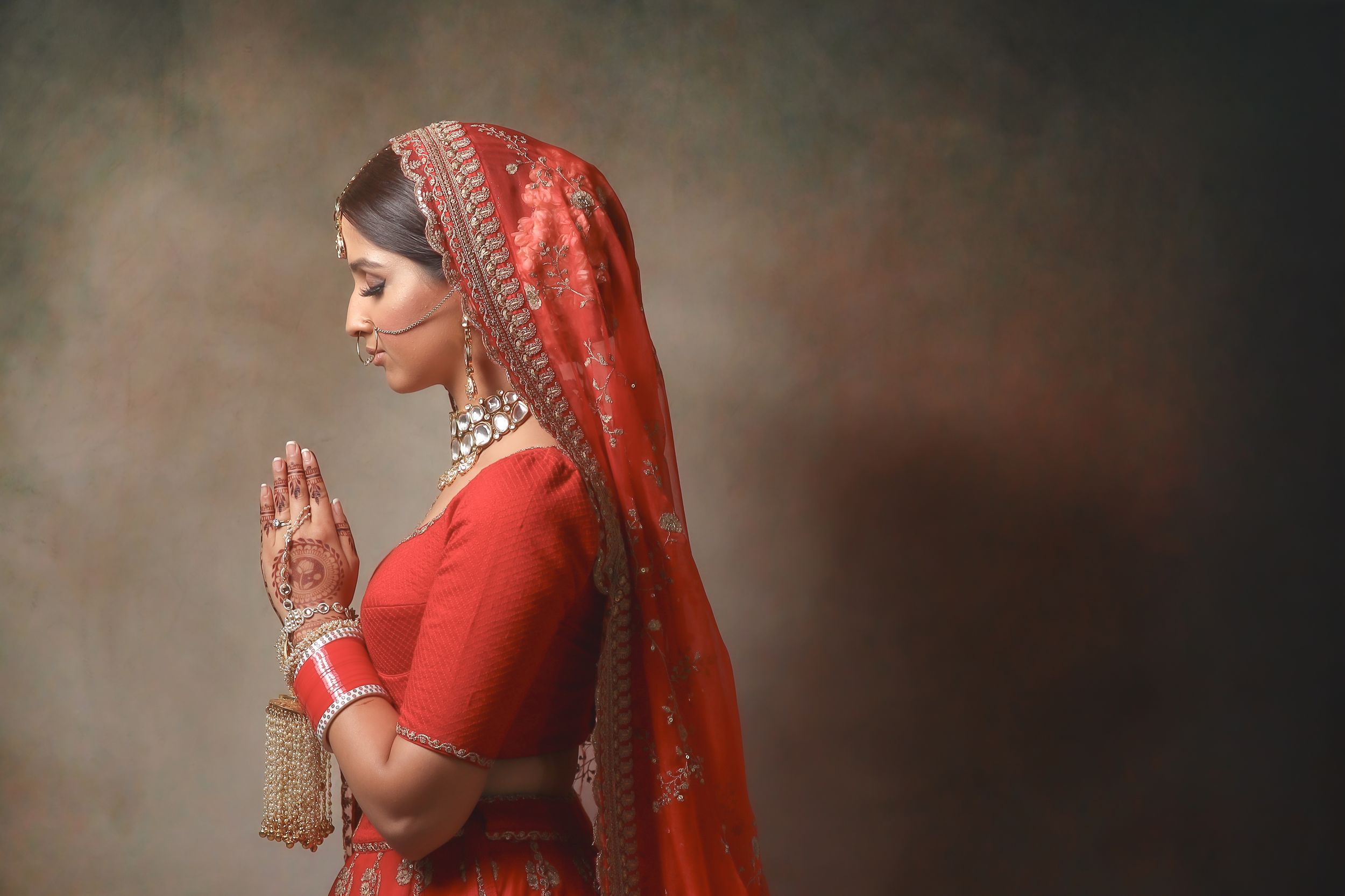 Indian bride in red saree with mehndi hands in prayer pose, wearing traditional gold jewelry and dupatta.