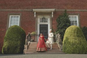 Indian bride and groom in traditional red and white attire holding hands on steps of elegant brick mansion.