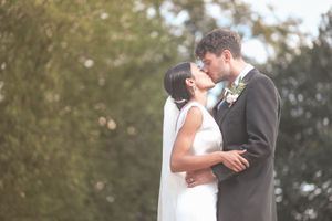 A bride and groom share a romantic kiss outdoors on their wedding day, surrounded by lush green trees.