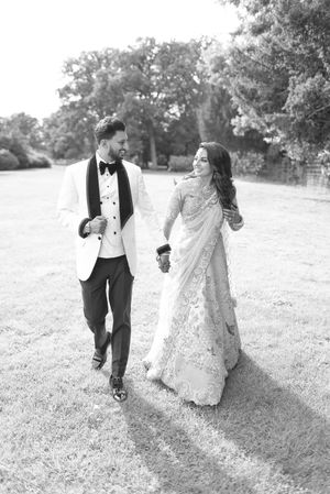 Black and white photo of a bride in a saree and groom in a tuxedo walking hand in hand on a lawn.