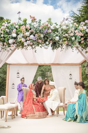 Indian wedding ceremony under a floral mandap with bride in red lehenga and groom in white sherwani.