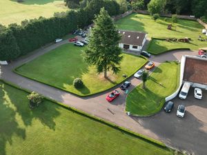 Aerial view of a countryside property with lush green lawns, a driveway, parked cars, and surrounding trees.