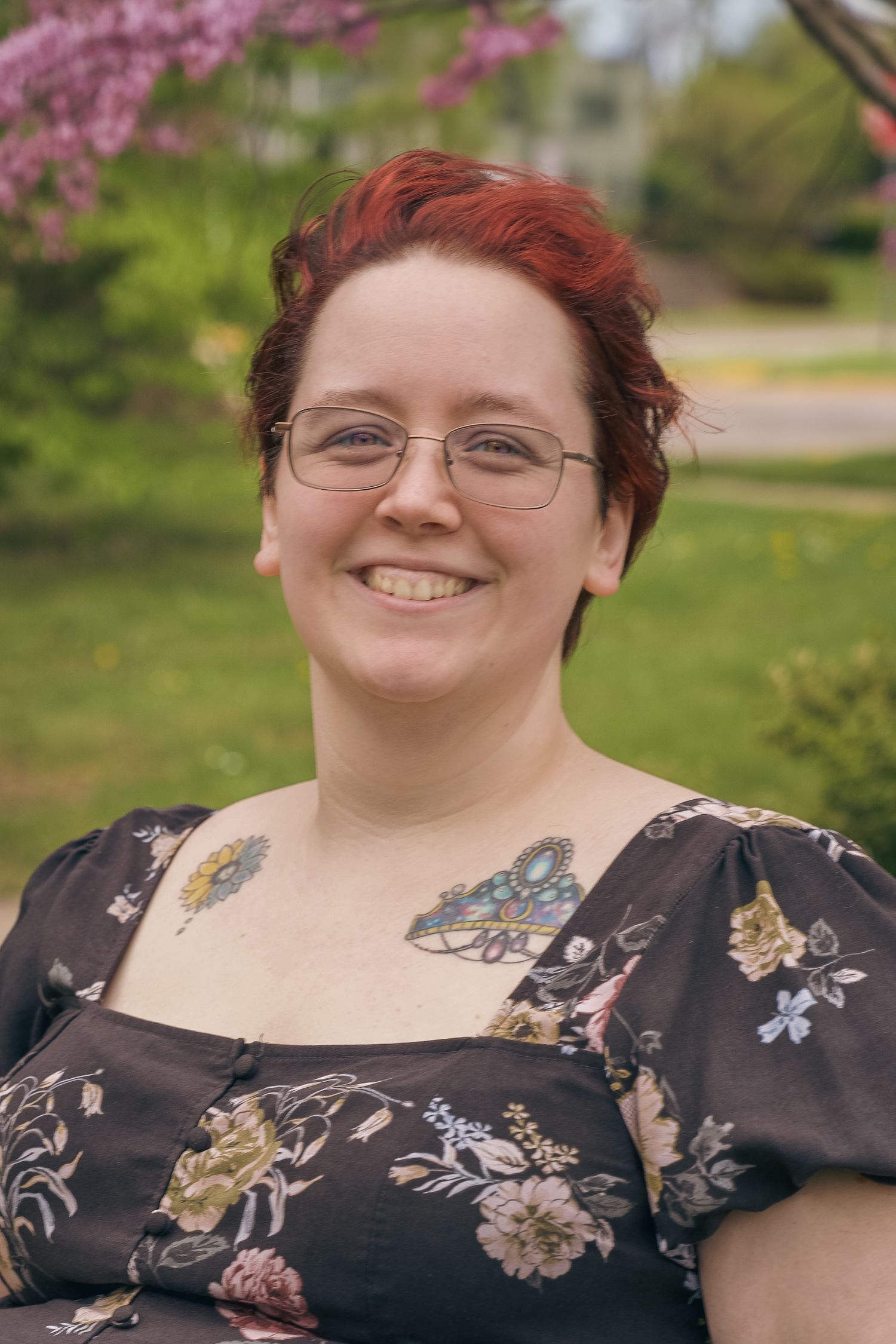 Smiling woman with red hair, glasses, and floral tattoos wearing a dark floral dress outdoors near blooming trees.