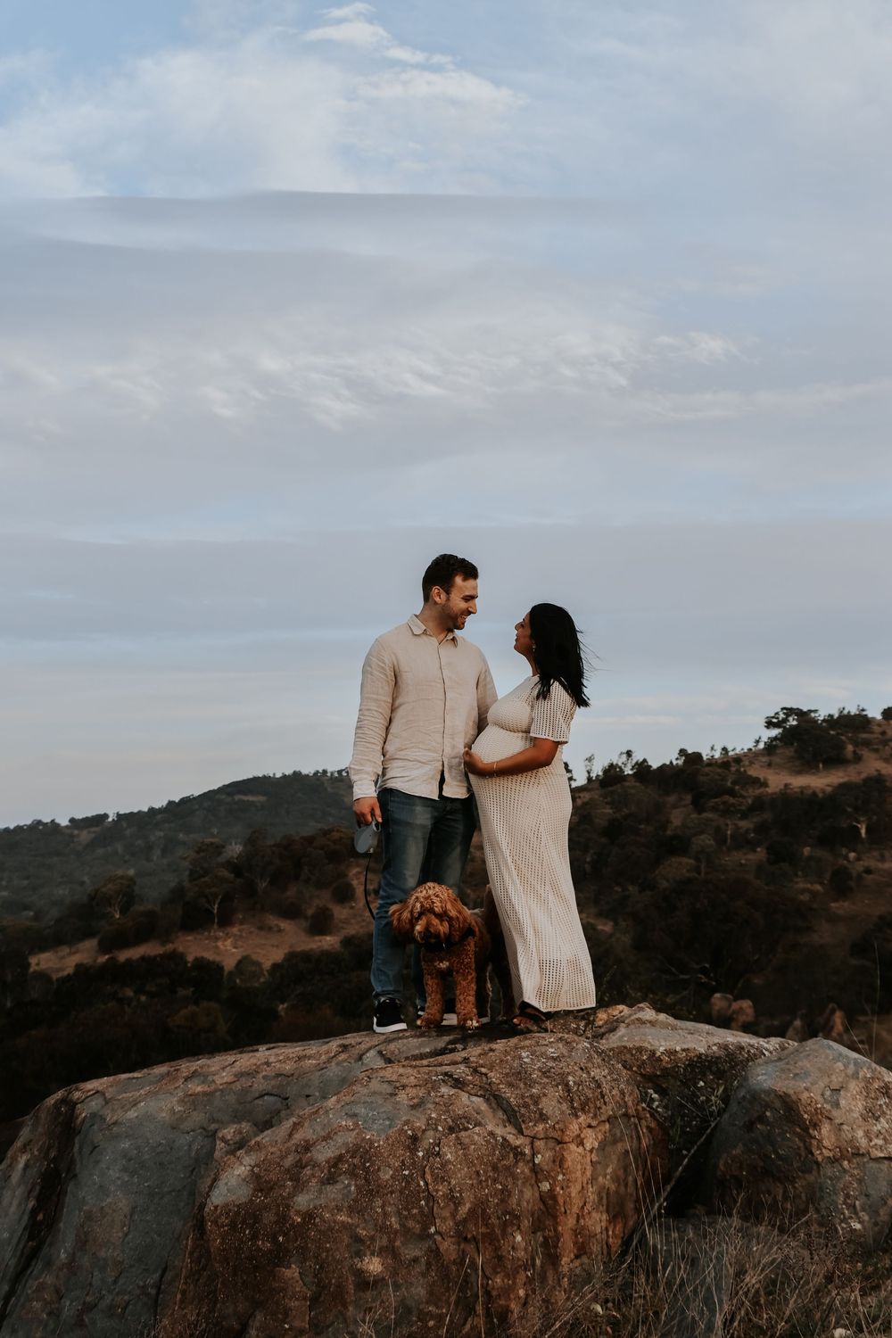 Couple standing on a rocky hilltop with their golden doodle dog, woman in white maternity dress, scenic mountain backdrop.