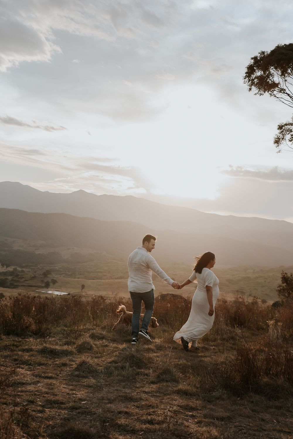Couple holding hands walking on a golden hilltop at sunset with mountain valley views in the background.